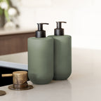 Close-up of two Smith & Co. sage green pump bottles with black dispensers positioned beside a brass tap on a white countertop.