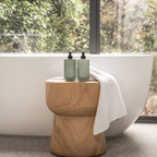 Two Smith & Co. sage green pump bottles displayed on a wooden stool beside a freestanding bathtub with a white towel draped over the stool.