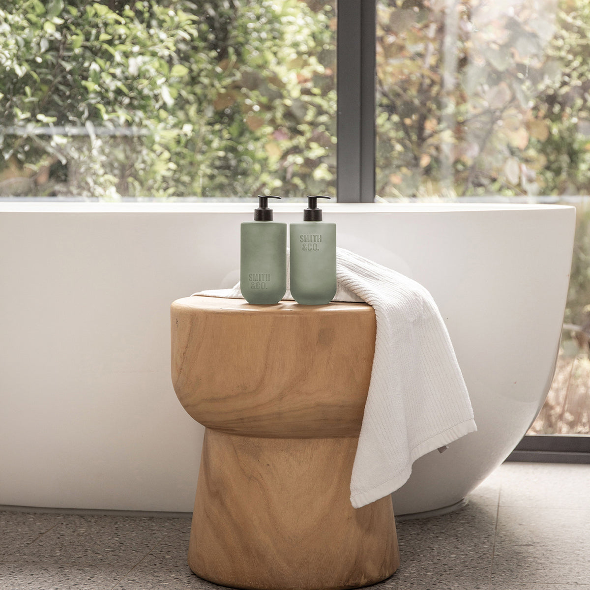 Two Smith & Co. sage green pump bottles displayed on a wooden stool beside a freestanding bathtub with a white towel draped over the stool.