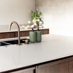 Two Smith & Co. sage green pump bottles displayed beside a brass tap on a white kitchen countertop with flowers in the background.