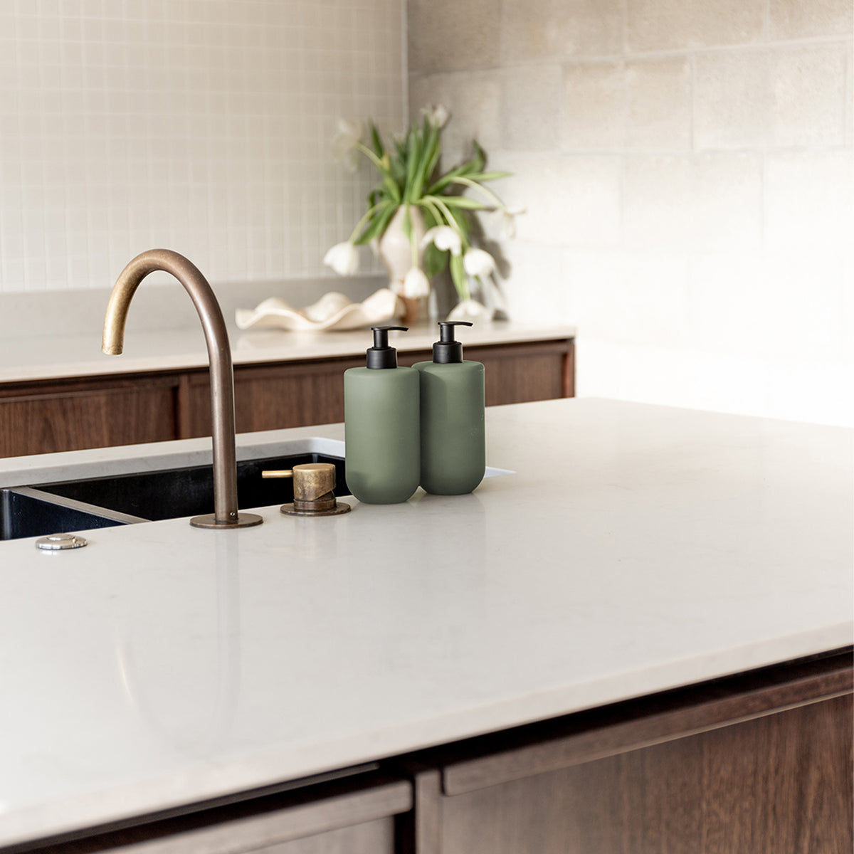 Two Smith & Co. sage green pump bottles displayed beside a brass tap on a white kitchen countertop with flowers in the background.
