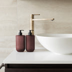 Two Smith & Co. deep burgundy pump bottles displayed beside a white vessel basin and brushed brass tap on a modern bathroom countertop.