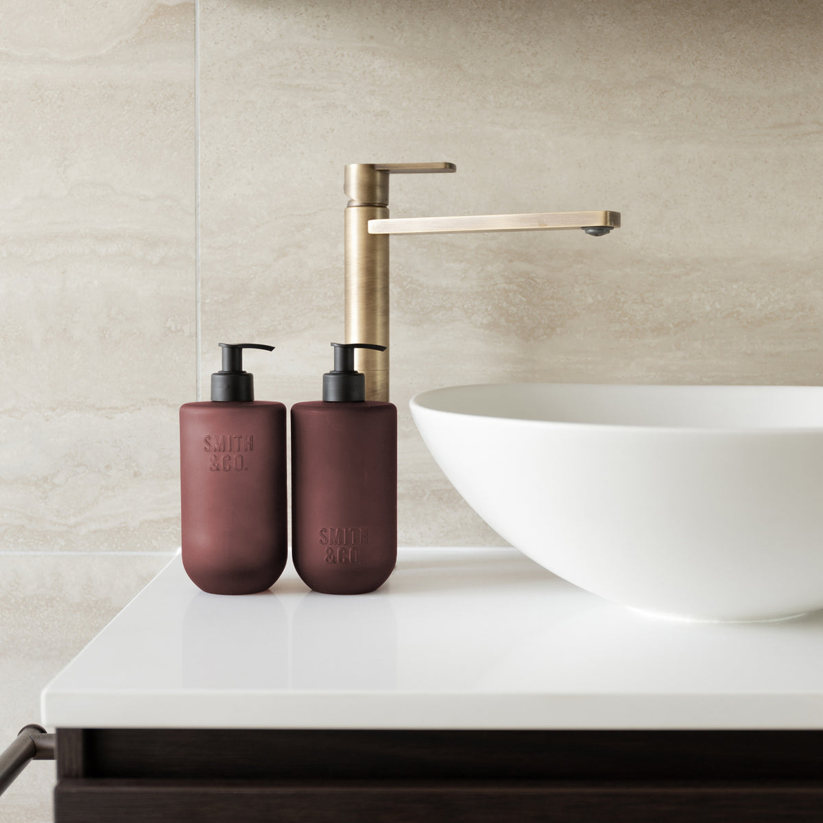 Two Smith & Co. deep burgundy pump bottles displayed beside a white vessel basin and brushed brass tap on a modern bathroom countertop.