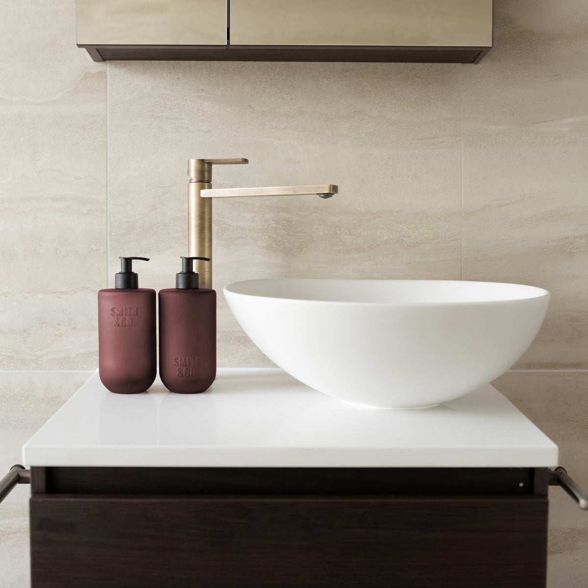 Two Smith & Co. deep burgundy pump bottles displayed beside a white vessel basin and brushed brass tap on a modern bathroom countertop.