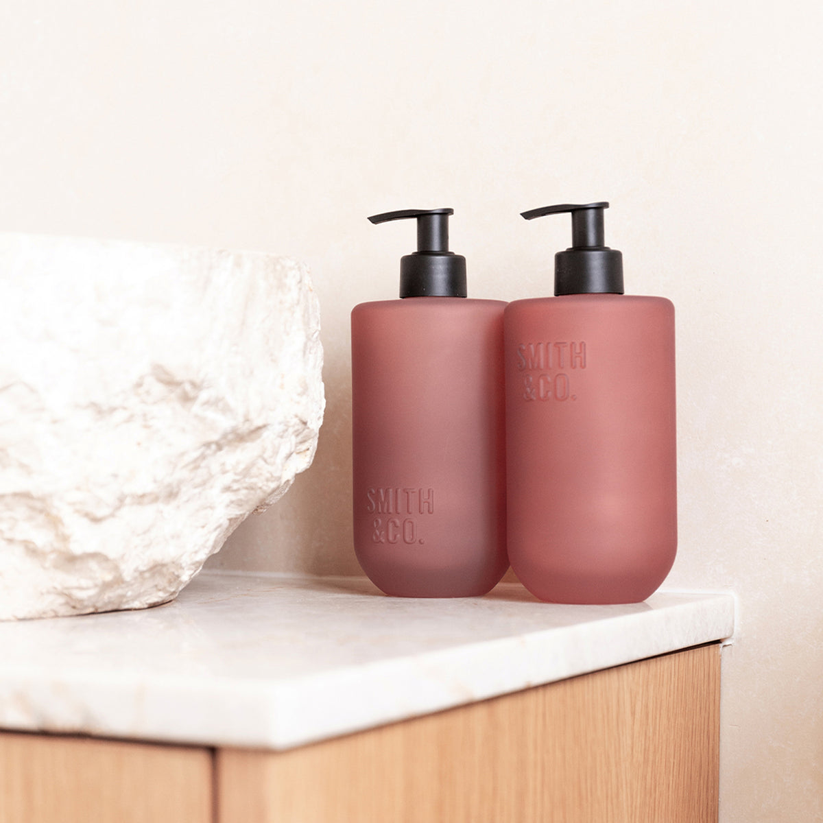 Close-up of two Smith & Co. Blush Rose & Blackberry pink pump bottles beside a white stone basin on a marble countertop.