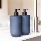 Close-up of two Smith & Co. Smoked Suede & Bergamot blue pump bottles with black dispensers beside a stainless steel kitchen tap.