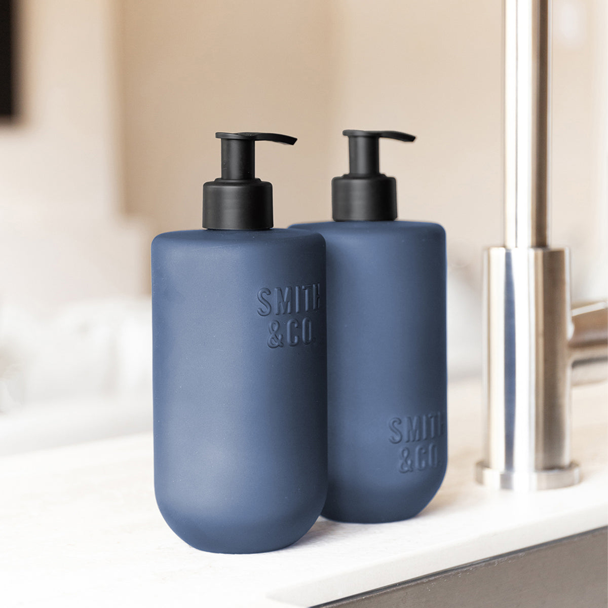 Close-up of two Smith & Co. Smoked Suede & Bergamot blue pump bottles with black dispensers beside a stainless steel kitchen tap.