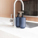 Close-up of two Smith & Co. Smoked Suede & Bergamot blue pump bottles with black dispensers beside a stainless steel kitchen tap.
