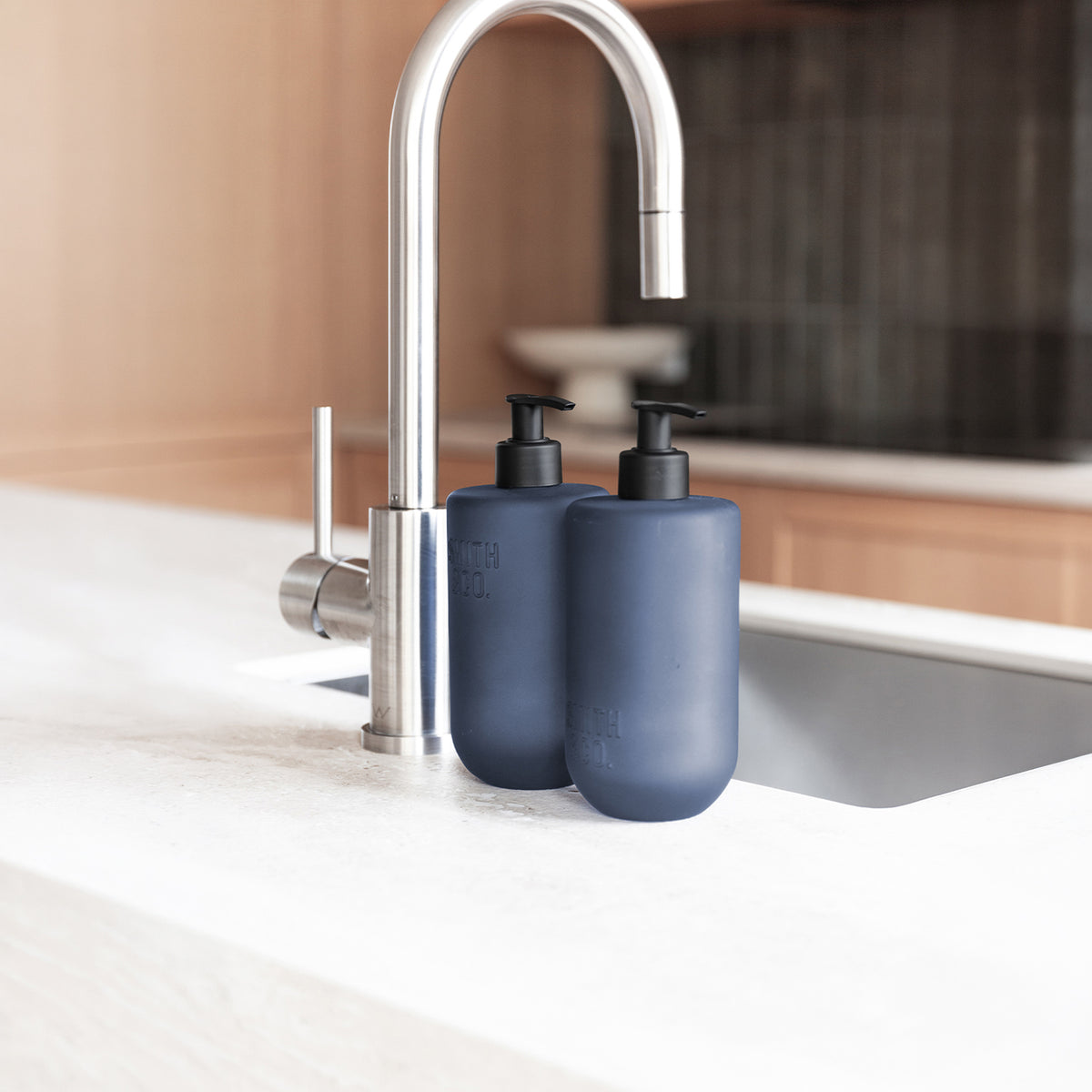Close-up of two Smith & Co. Smoked Suede & Bergamot blue pump bottles with black dispensers beside a stainless steel kitchen tap.