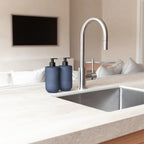 Two Smith & Co. Smoked Suede & Bergamot blue pump bottles positioned beside a stainless steel sink and curved tap in a modern kitchen.