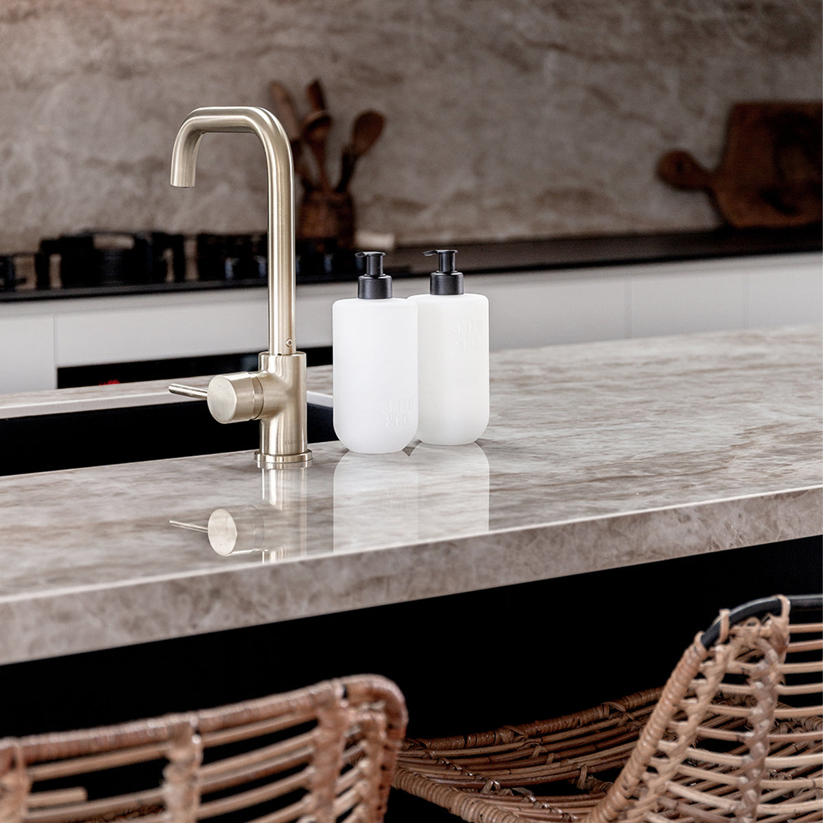 Two Smith & Co. Tonka & White Musk white pump bottles displayed beside a brushed brass tap on a marble kitchen island with woven bar stools.