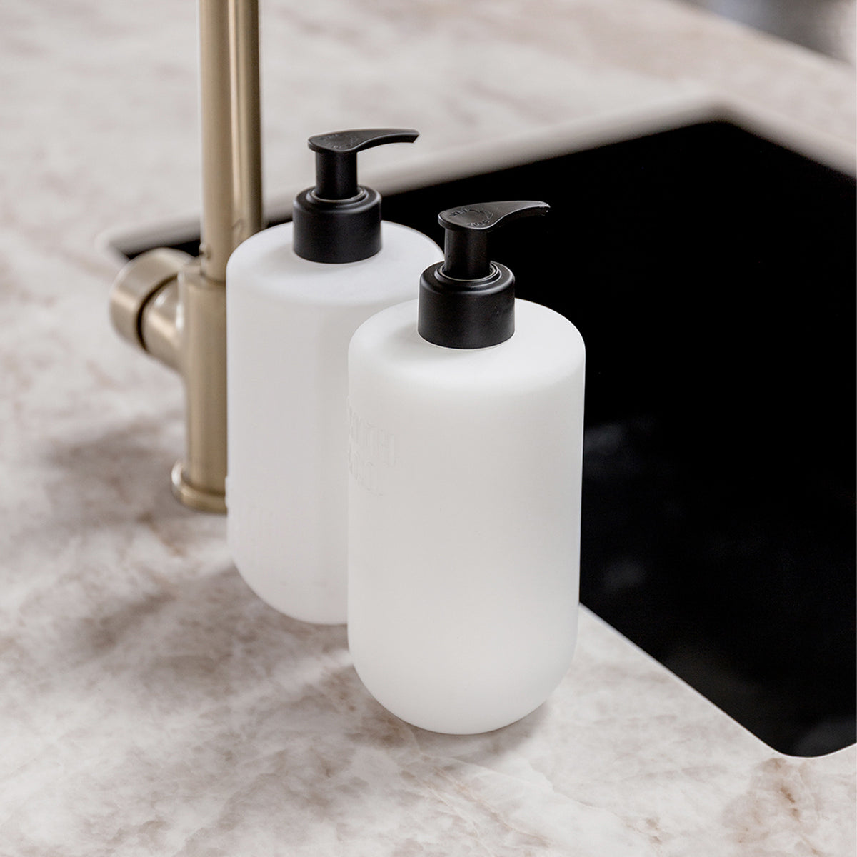 Close-up of two Smith & Co. Tonka & White Musk white pump bottles beside a brass kitchen tap and black sink on a marble countertop.