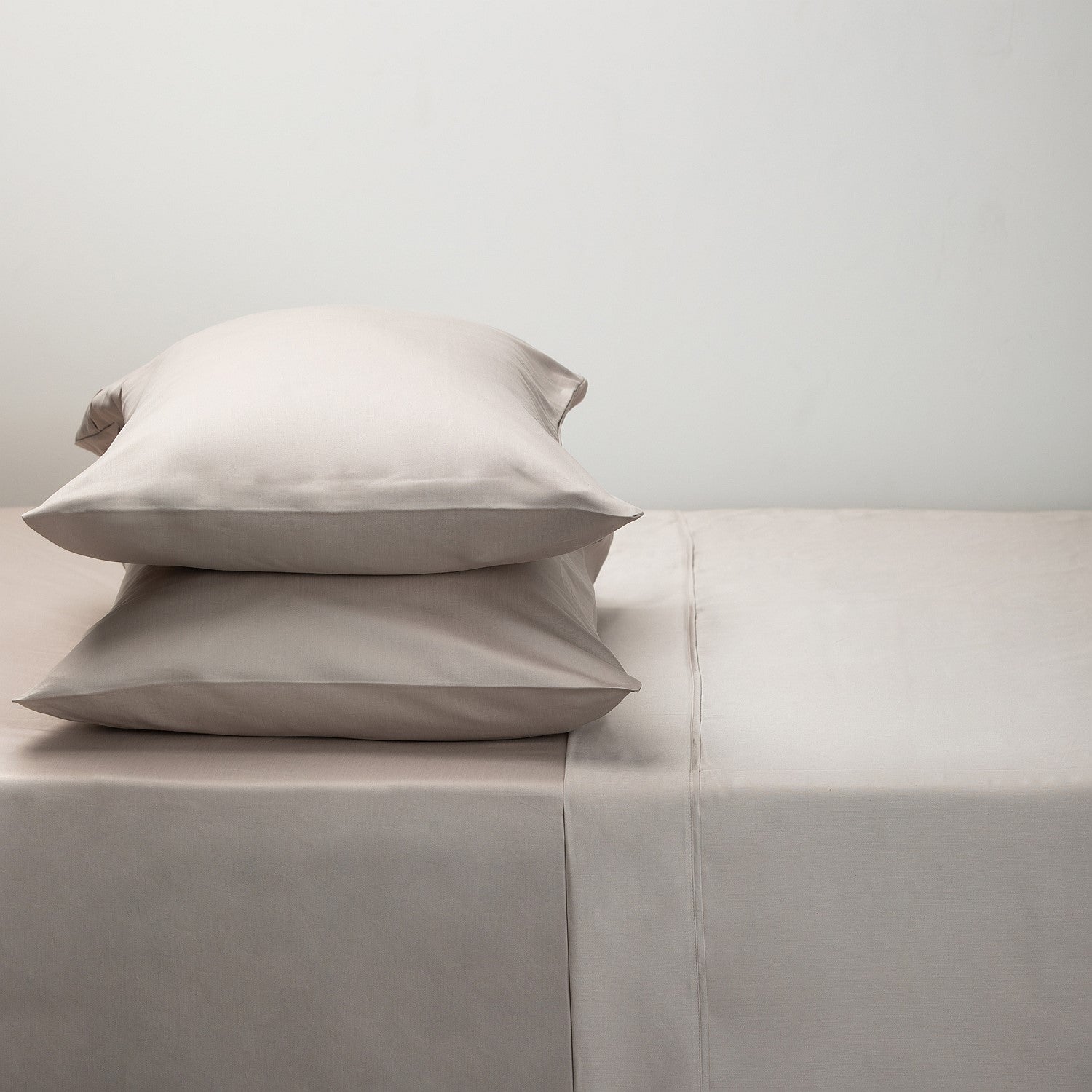 Two pillows stacked on a bed with light gray bedding against a plain background