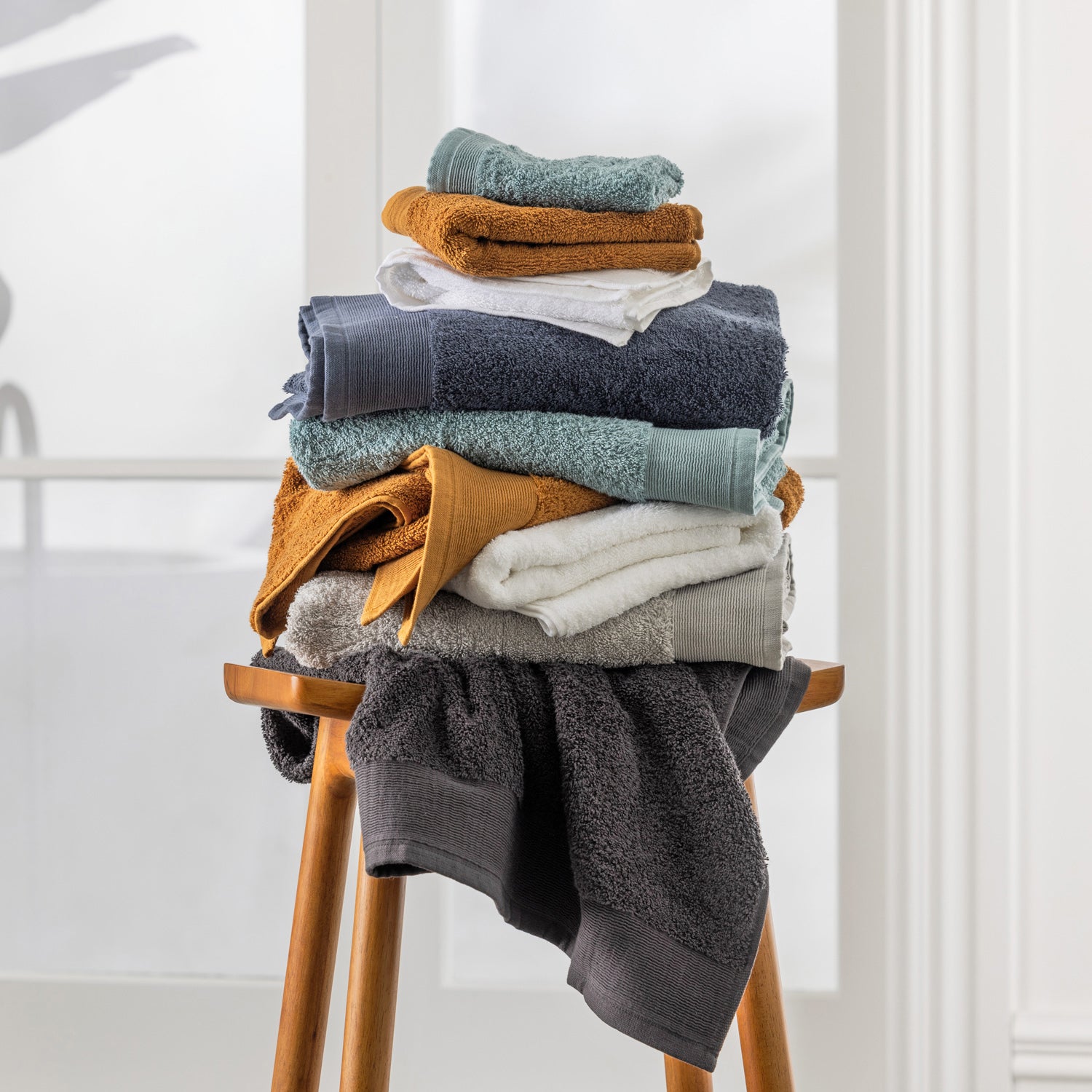 Stack of Egyptian cotton face cloths in assorted colours displayed on a wooden stool in a bright bathroom setting.