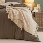 Home Co. Mateo Throw in Natural draped over the end of a bed, showcasing the soft woven texture and tassel edging in a warm, neutral bedroom setting.