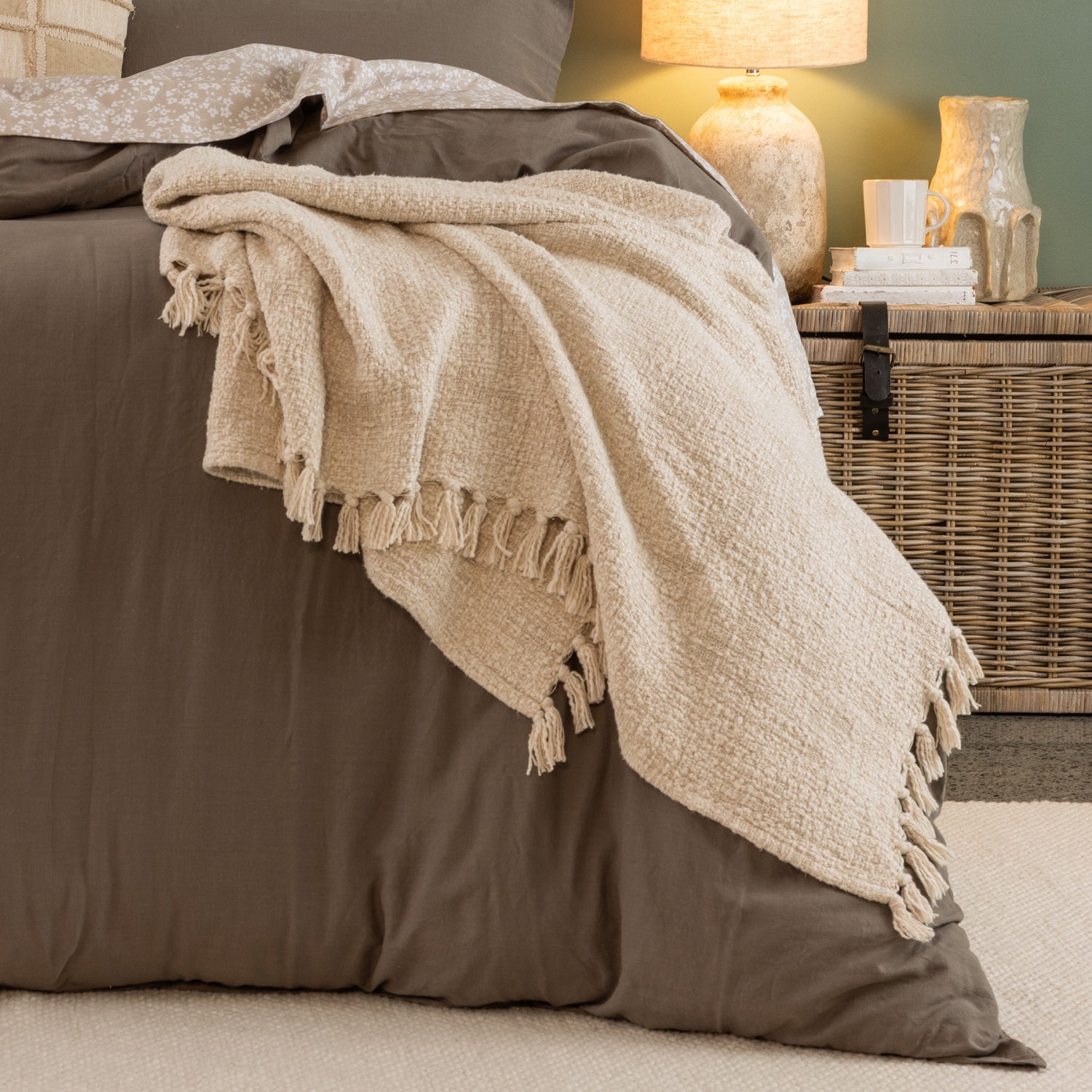 Home Co. Mateo Throw in Natural draped over the end of a bed, showcasing the soft woven texture and tassel edging in a warm, neutral bedroom setting.