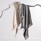 Home Co. Mateo Throws in Natural and Silver draped over a natural driftwood branch, showcasing the textured woven design and decorative tassel fringe.
