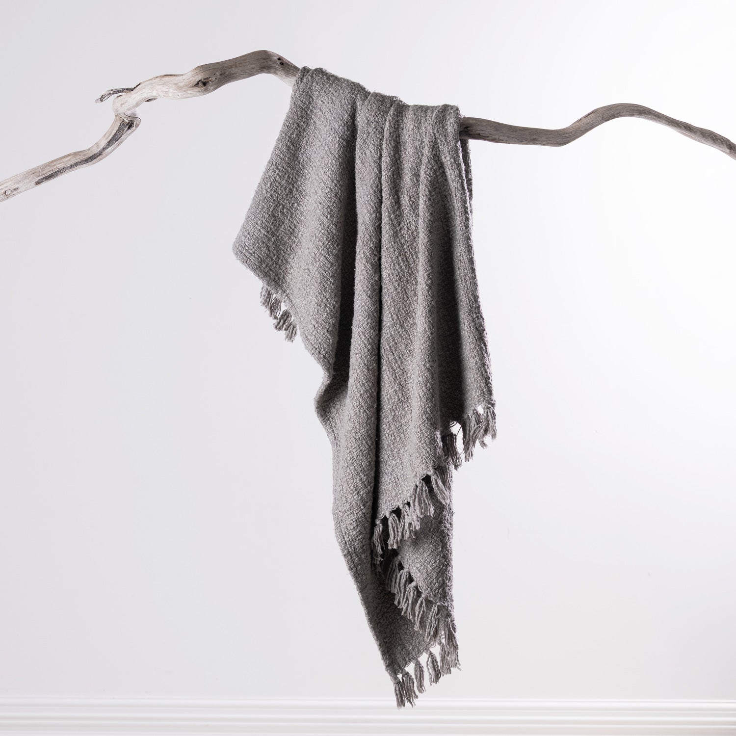 Home Co. Mateo Throw in Silver draped over a natural driftwood branch, featuring a textured woven finish with tassel edging.