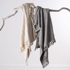 Home Co. Mateo Throws in Natural and Silver draped over a natural driftwood branch, showcasing the textured woven design and decorative tassel fringe.