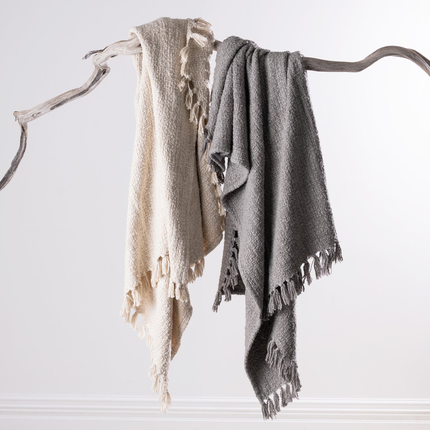 Home Co. Mateo Throws in Natural and Silver draped over a natural driftwood branch, showcasing the textured woven design and decorative tassel fringe.
