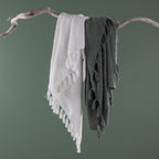 White and sage green textured throw blankets with tassel fringe draped together over a natural driftwood branch against a sage green background.