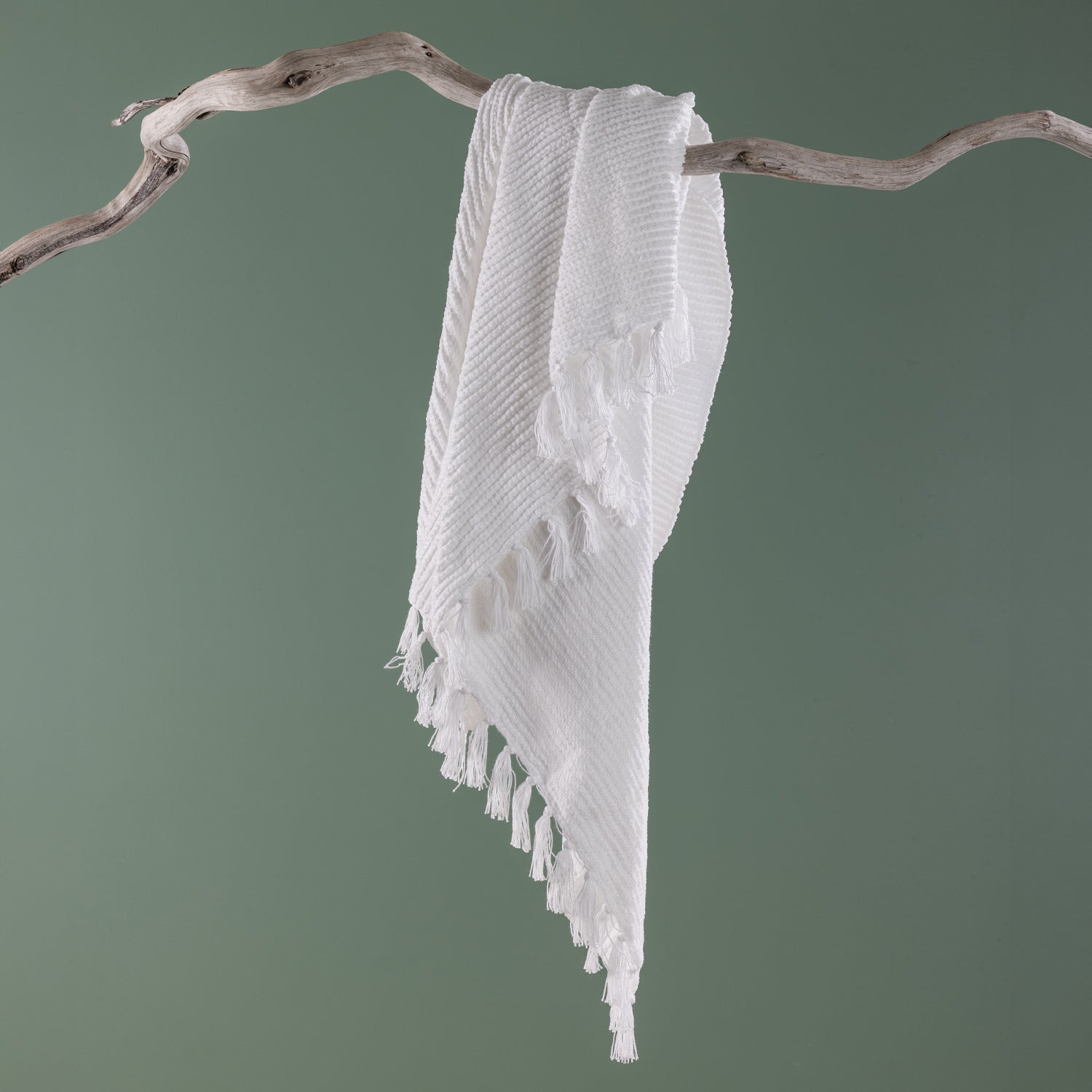 White textured throw blanket with tassel fringe draped over a natural driftwood branch against a soft sage green background.