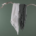 White and sage green textured throw blankets with tassel fringe draped together over a natural driftwood branch against a sage green background.