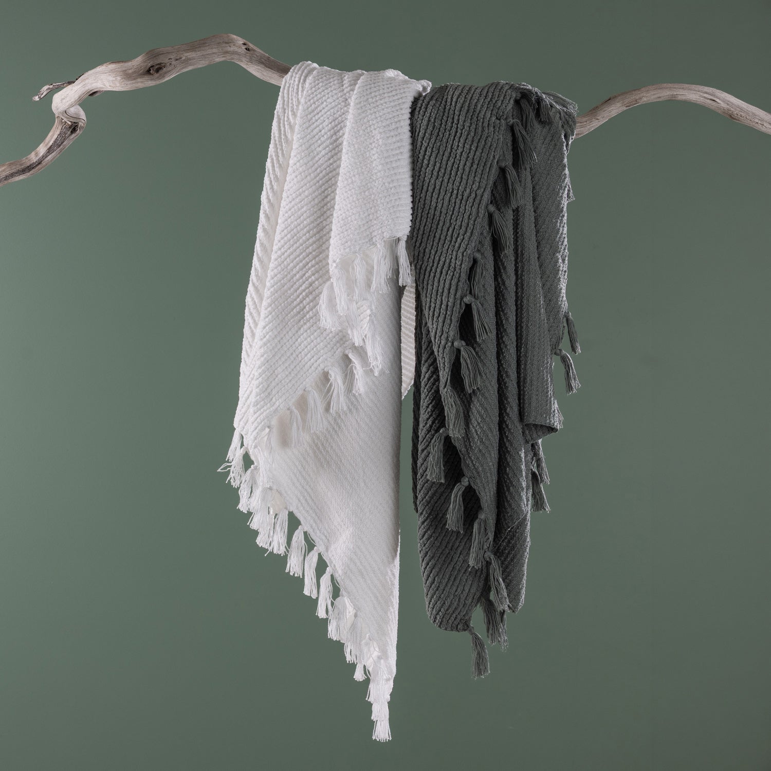 White and sage green textured throw blankets with tassel fringe draped together over a natural driftwood branch against a sage green background.