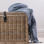 A textured blue woven throw with fringe detailing is casually draped over a natural wicker storage basket, creating a relaxed and inviting home styling display.