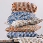 A coordinated stack of textured woven cushions and fringed throws in warm rust, natural, and blue tones creates a cosy and inviting home décor display.