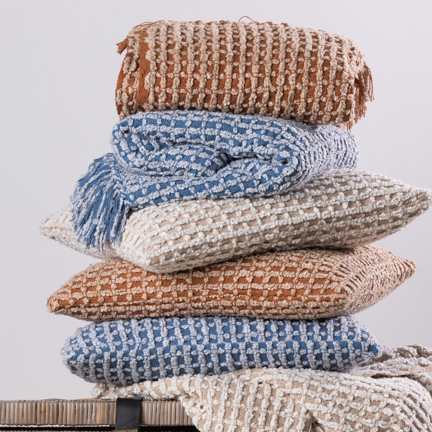 A coordinated stack of textured woven cushions and fringed throws in warm rust, natural, and blue tones creates a cosy and inviting home décor display.