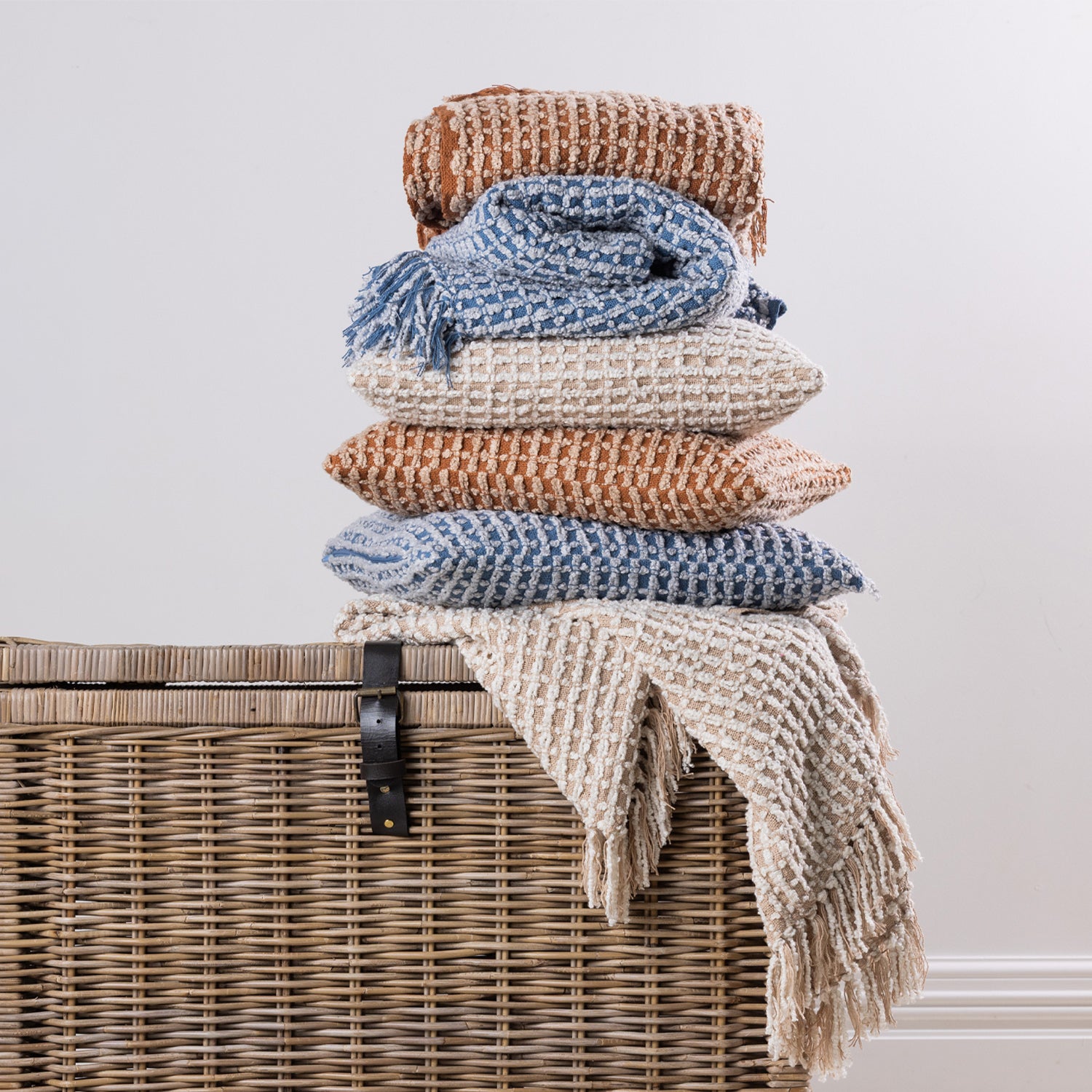 A coordinated stack of textured woven cushions and fringed throws in warm rust, natural, and blue tones creates a cosy and inviting home décor display.