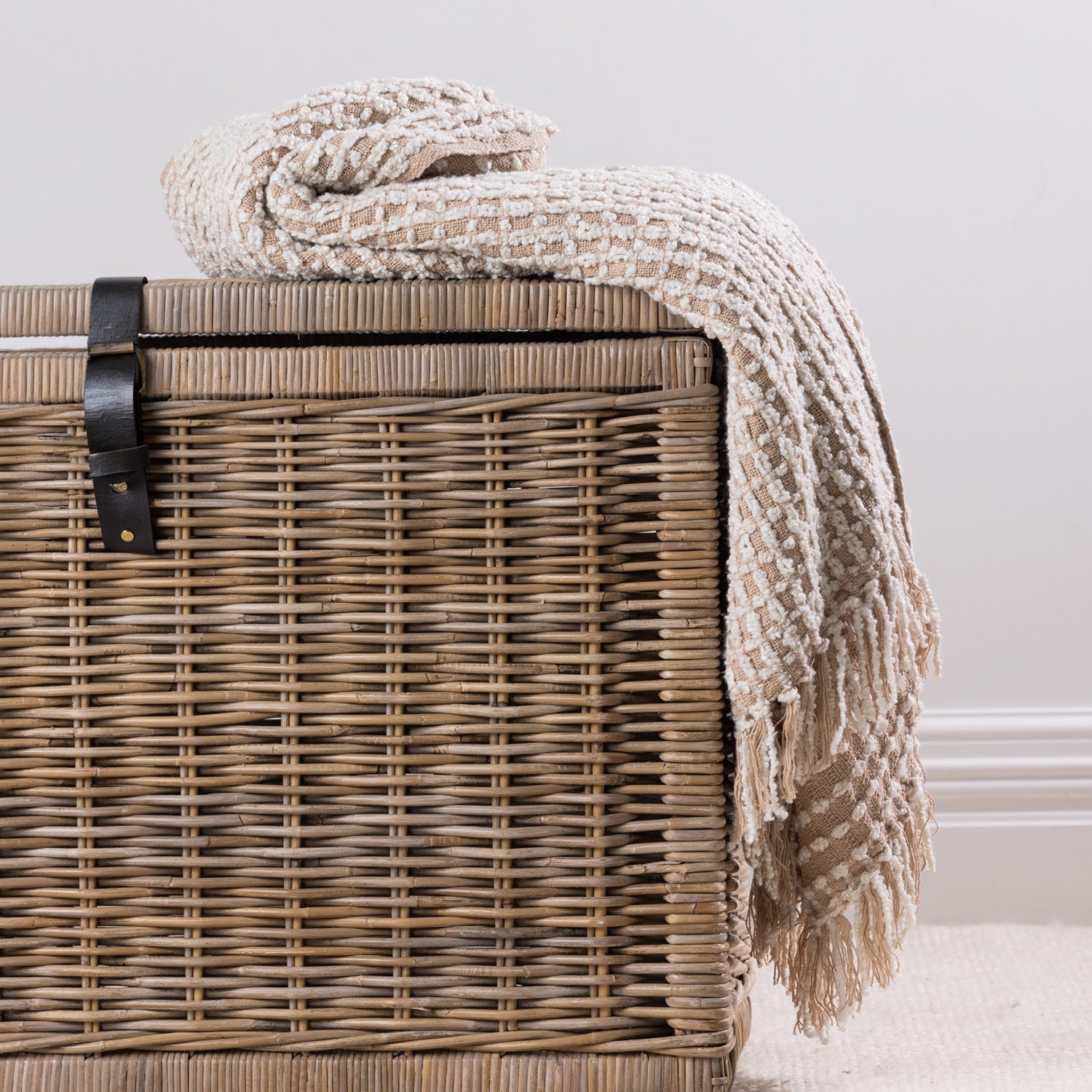 A textured natural woven throw with fringe detailing is casually draped over a natural wicker storage basket, creating a relaxed and inviting home styling display.