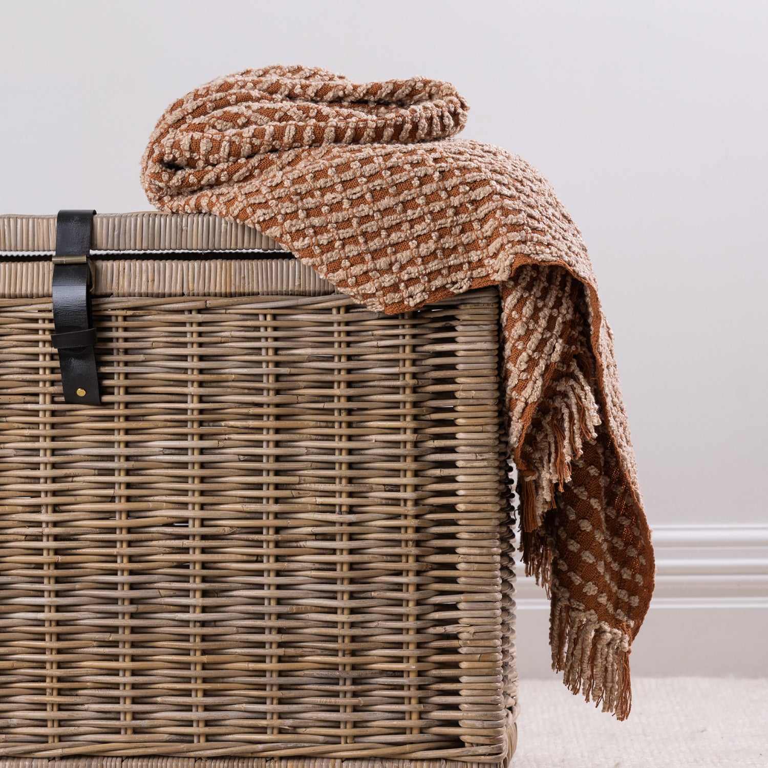 A textured rust woven throw with fringe detailing is casually draped over a natural wicker storage basket, creating a relaxed and inviting home styling display.