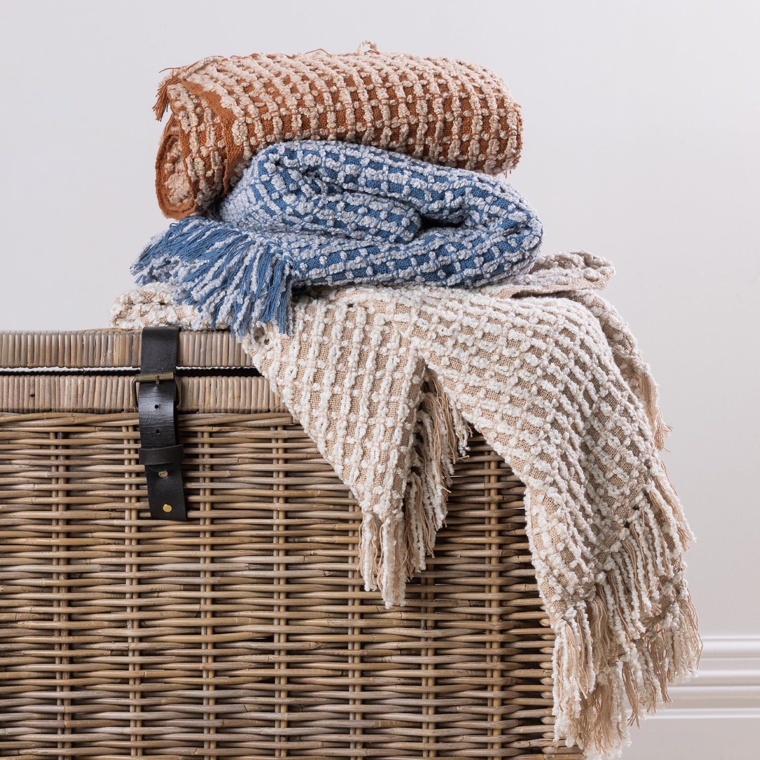 A coordinated stack of textured woven  fringed throws in warm rust, natural, and blue tones creates a cosy and inviting home décor display.