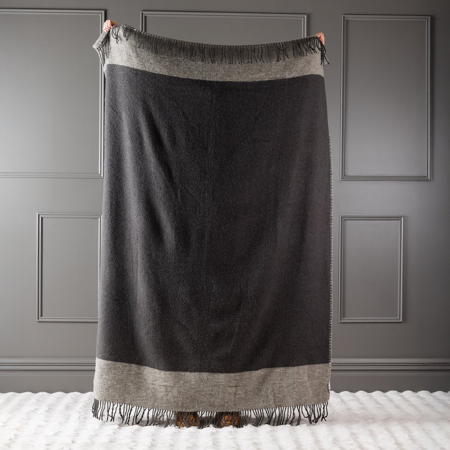 Charcoal New Zealand wool throw held up against a grey panelled wall.