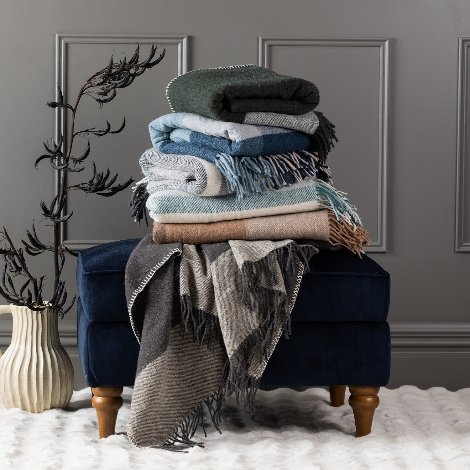 Stack of New Zealand wool throws in assorted colours styled on a navy ottoman with a decorative vase.