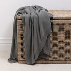 A soft knitted cotton throw in a charcoal tone is casually draped over a woven wicker storage trunk, adding inviting texture and effortless style to the space.