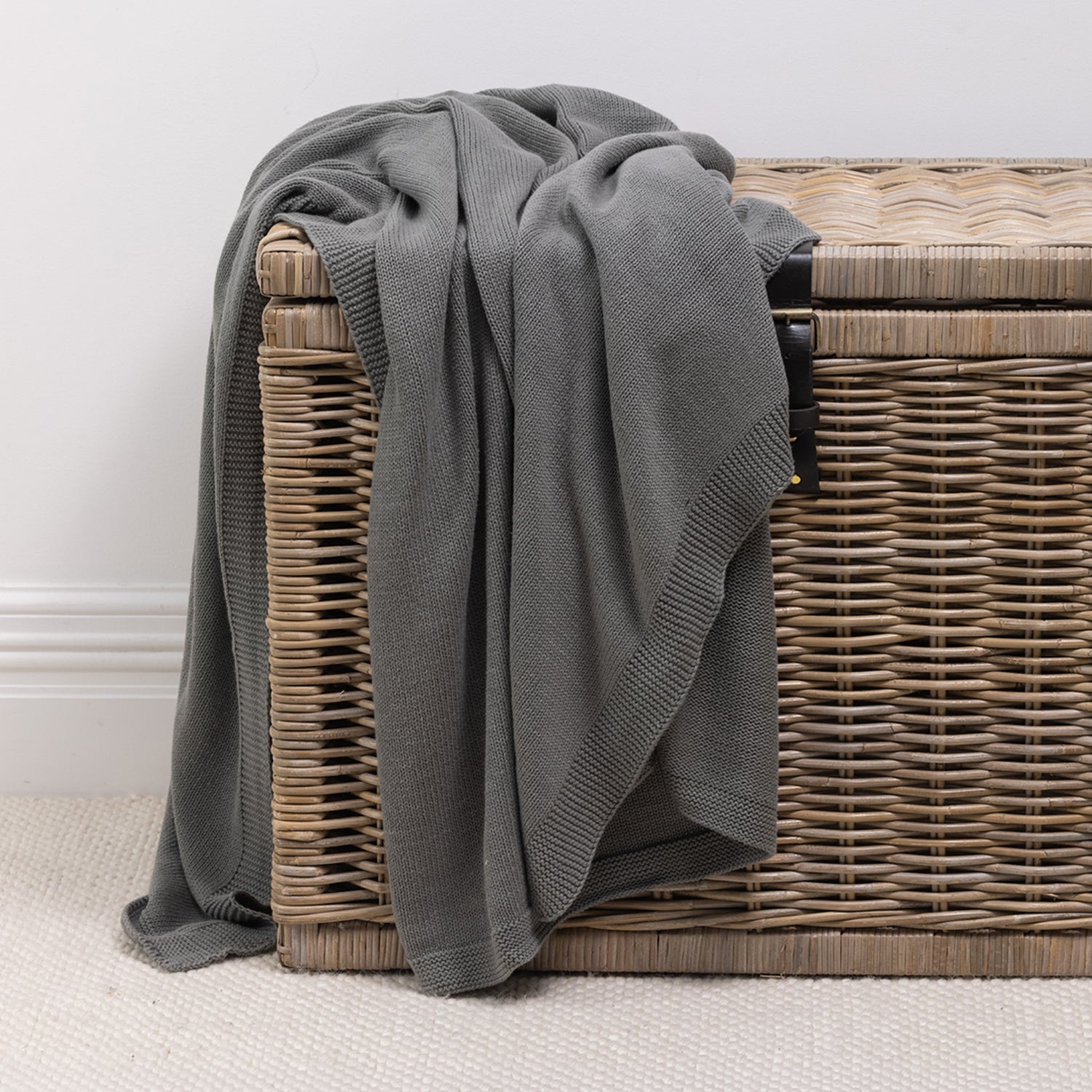 A soft knitted cotton throw in a charcoal tone is casually draped over a woven wicker storage trunk, adding inviting texture and effortless style to the space.