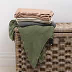 A collection of knitted cotton throws in soft, versatile colours is neatly folded and styled over a woven wicker storage trunk, bringing warmth, texture and effortless style to any living space.
