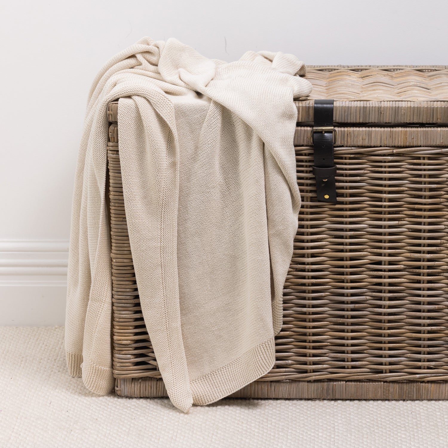 A soft knitted cotton throw in a neutral cream tone is casually draped over a woven wicker storage trunk, adding inviting texture and effortless style to the space.