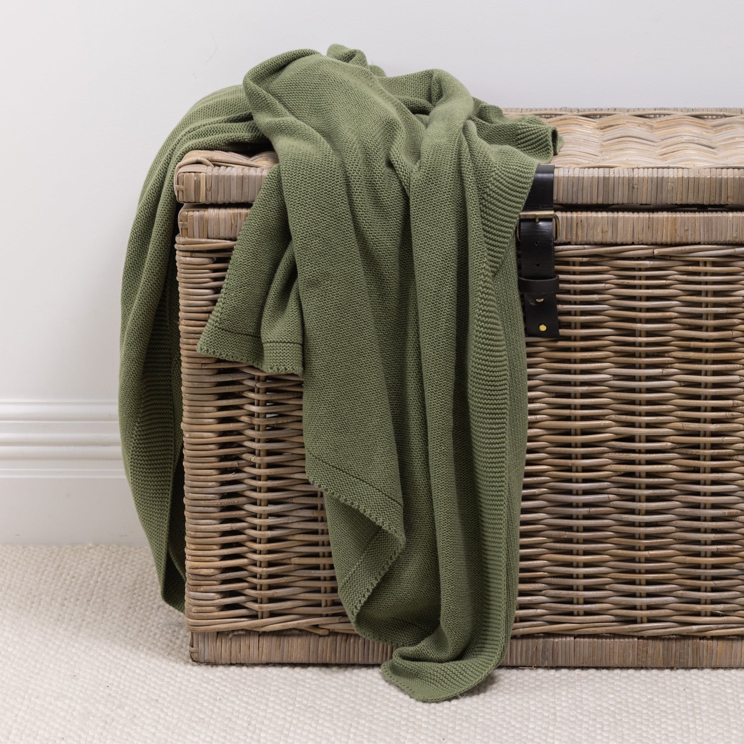 A soft knitted cotton throw in a forest green tone is casually draped over a woven wicker storage trunk, adding inviting texture and effortless style to the space.