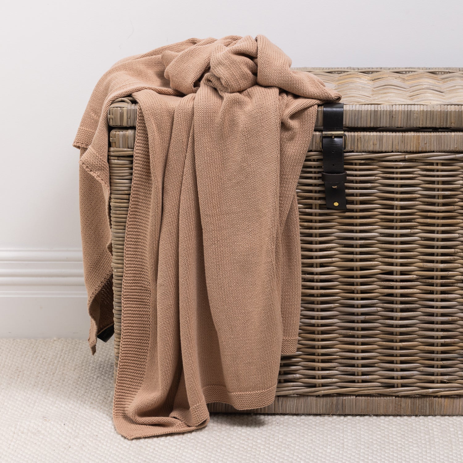 A soft knitted cotton throw in a warm natural clay is casually draped over a woven wicker storage trunk, adding inviting texture and effortless style to the space.