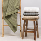 A textured cotton waffle throw is draped over a wooden ladder alongside neatly folded coordinating throws, showcasing its timeless design and effortless layering appeal.