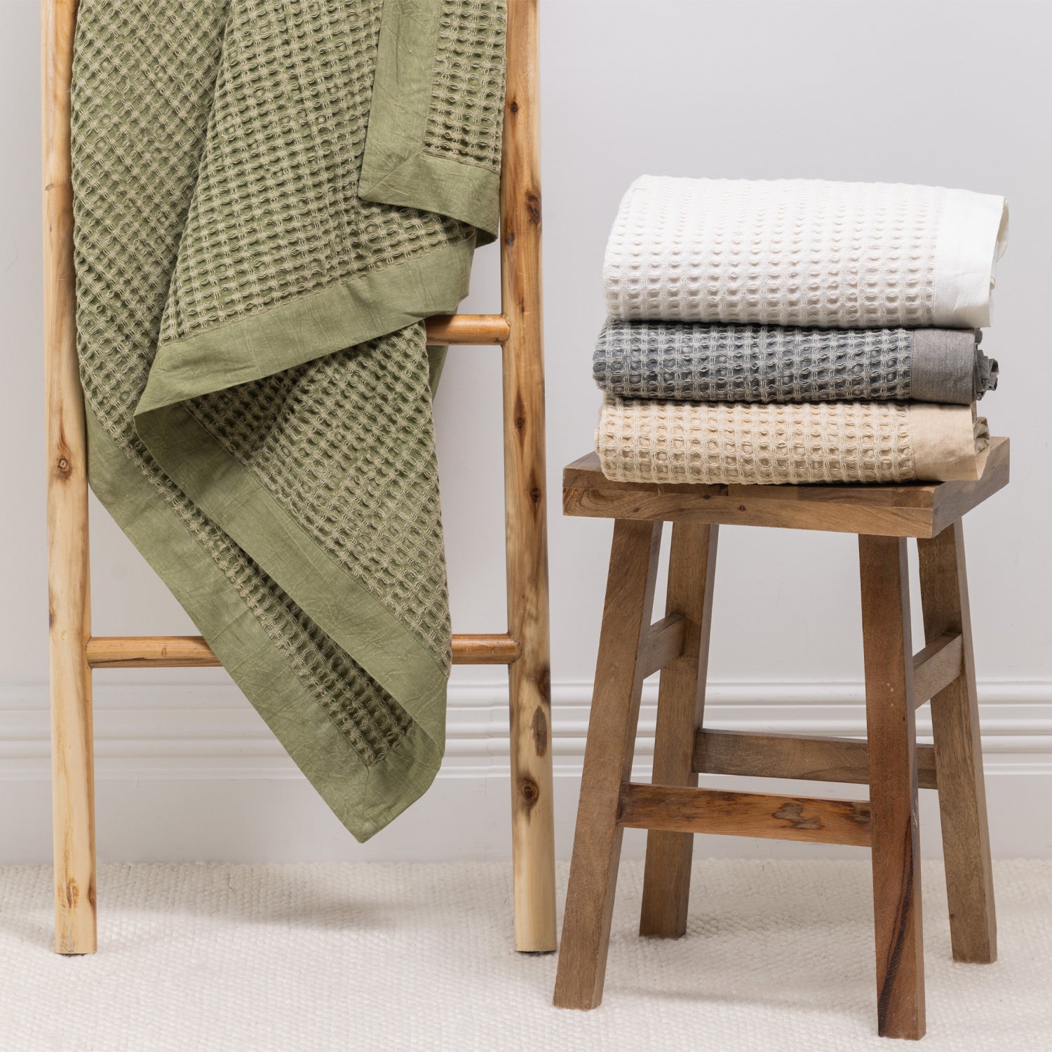 A textured cotton waffle throw is draped over a wooden ladder alongside neatly folded coordinating throws, showcasing its timeless design and effortless layering appeal.