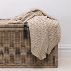A chunky knit throw in a natural color, draped over a brown woven wicker chest against a neutral light gray wall.