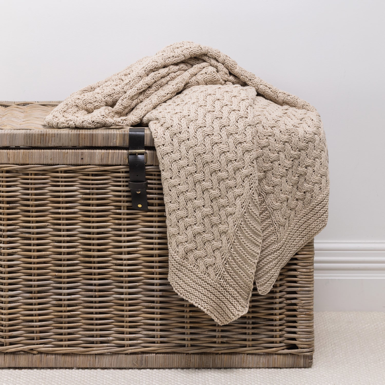 A chunky knit throw in a natural color, draped over a brown woven wicker chest against a neutral light gray wall.