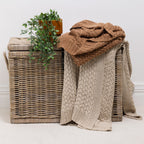 Two chunky knit throws draped over a woven wicker storage basket, with a small potted green plant on top. One throw is a rust and the other is a natural, set against a neutral white wall and light carpet.