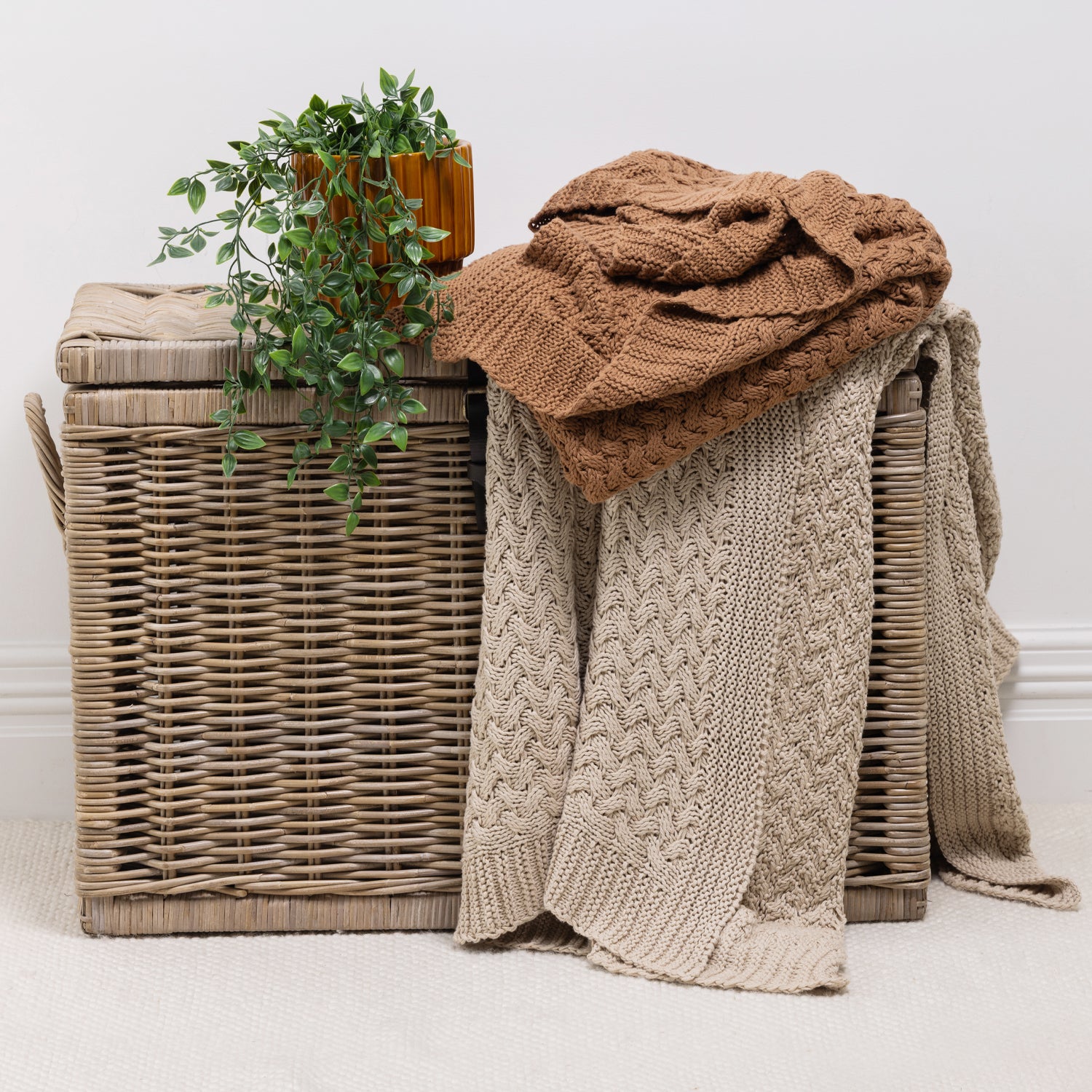 Two chunky knit throws draped over a woven wicker storage basket, with a small potted green plant on top. One throw is a rust and the other is a natural, set against a neutral white wall and light carpet.