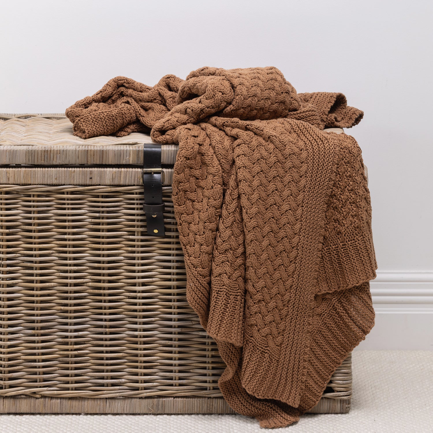 A chunky knit throw in a rust color, draped over a brown woven wicker chest against a neutral light gray wall.